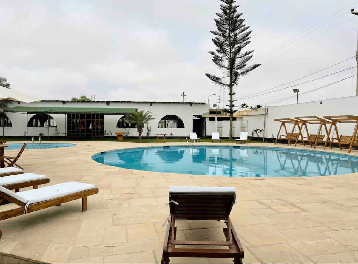 A large outdoor pool is surrounded by a spacious tiled patio. Loungers with white cushions are positioned for relaxation. In the background, a tiered palm tree adds a touch of greenery, along with a covered seating area visible through the open entrance.