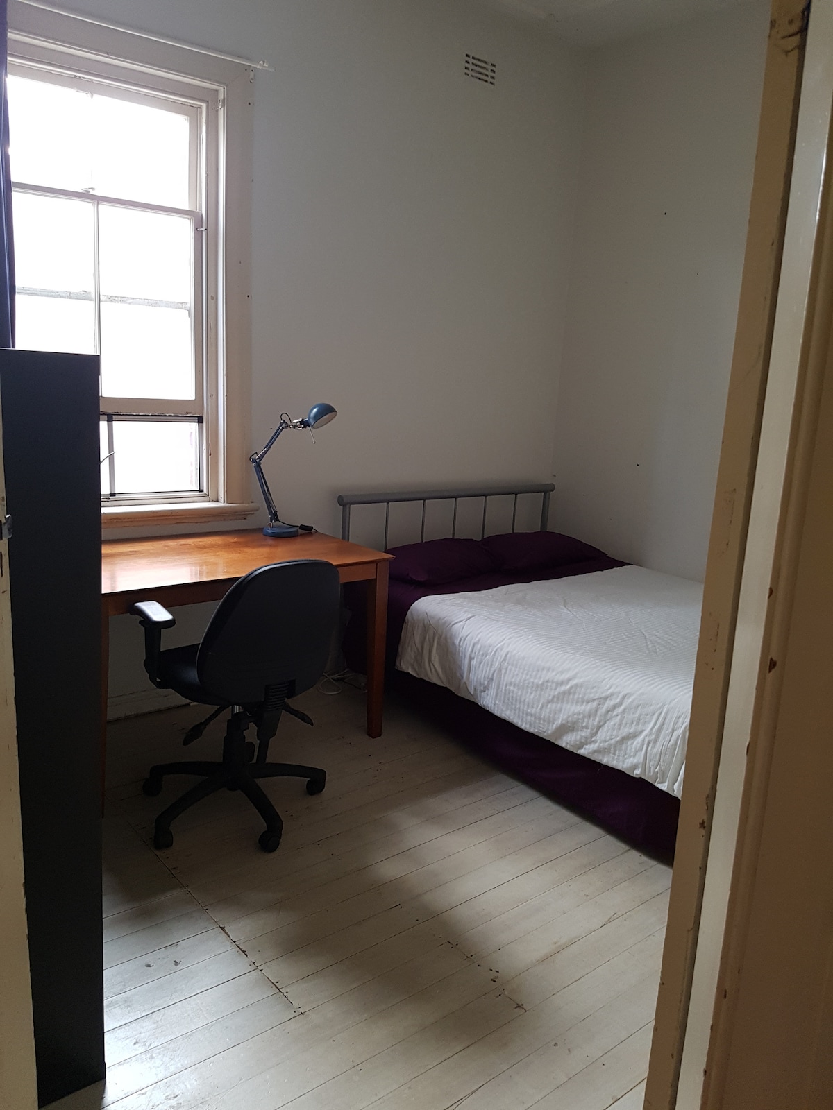 A simple room features a comfortable bed with a purple accent and a workspace with a wooden desk and a black office chair. Natural light enters through a large window, illuminating the light-colored floorboards and white walls.