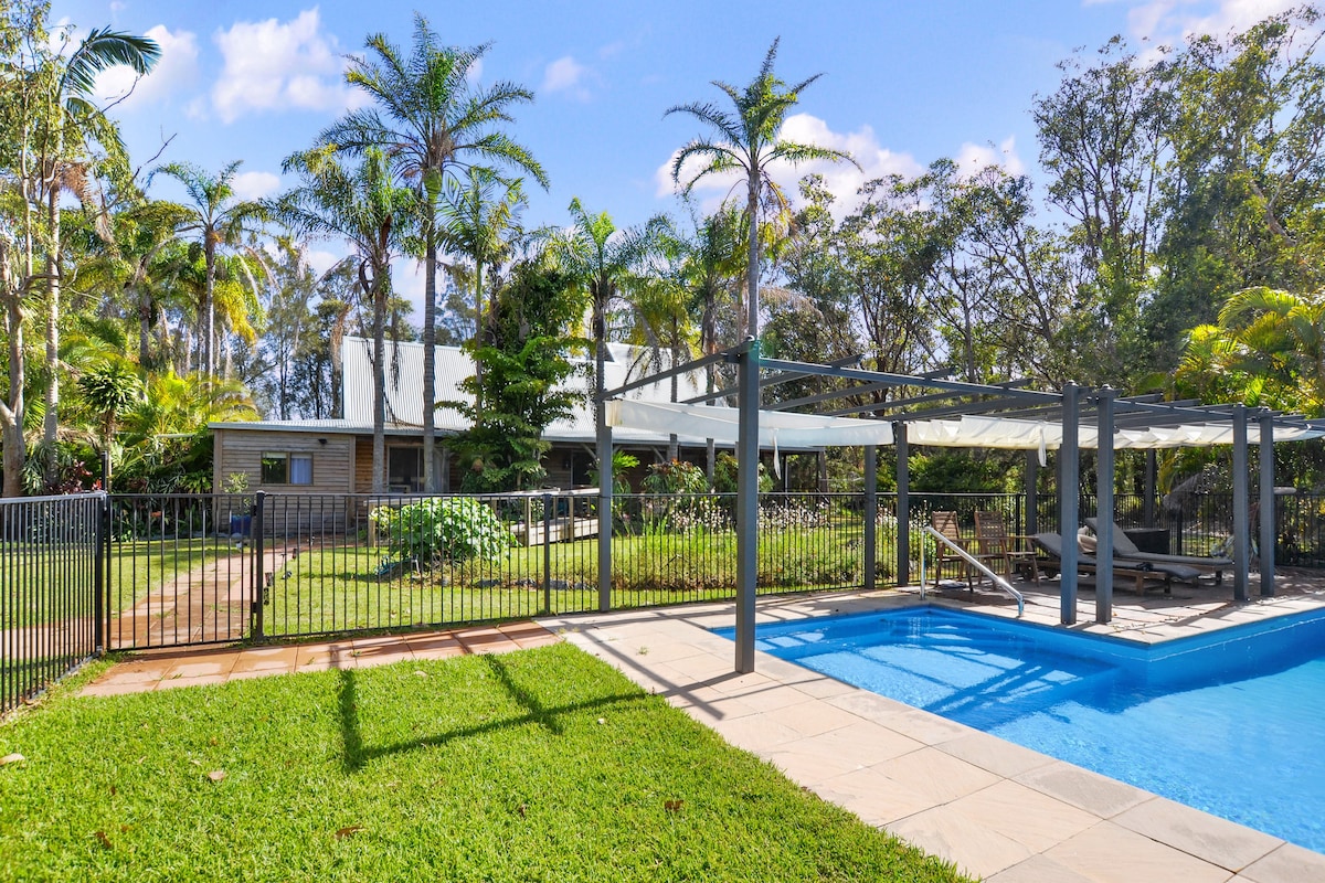 The image showcases a spacious outdoor area featuring a crystal-clear pool adjacent to a rustic cottage. Lush greenery surrounds the space, while palm trees add tropical charm. A shaded seating area is visible next to the pool, providing a relaxing ambiance for guests.