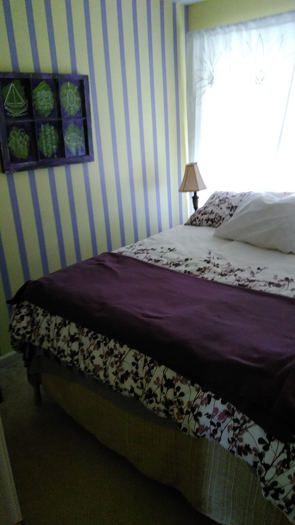 A cozy bedroom features soft, yellow and purple striped walls. A spacious bed is dressed in a floral-patterned comforter and white pillows. Natural light enters through a sheer window covering, and a small bedside lamp adds function and warmth. A framed artwork hangs on the wall.
