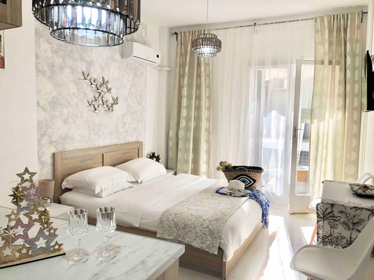 The studio apartment features a bright sleeping area with a spacious bed and soft linens. Natural light filters through large, sheer curtains. Decor elements include a decorative wall feature and modern light fixtures, while a small dining table is positioned nearby.