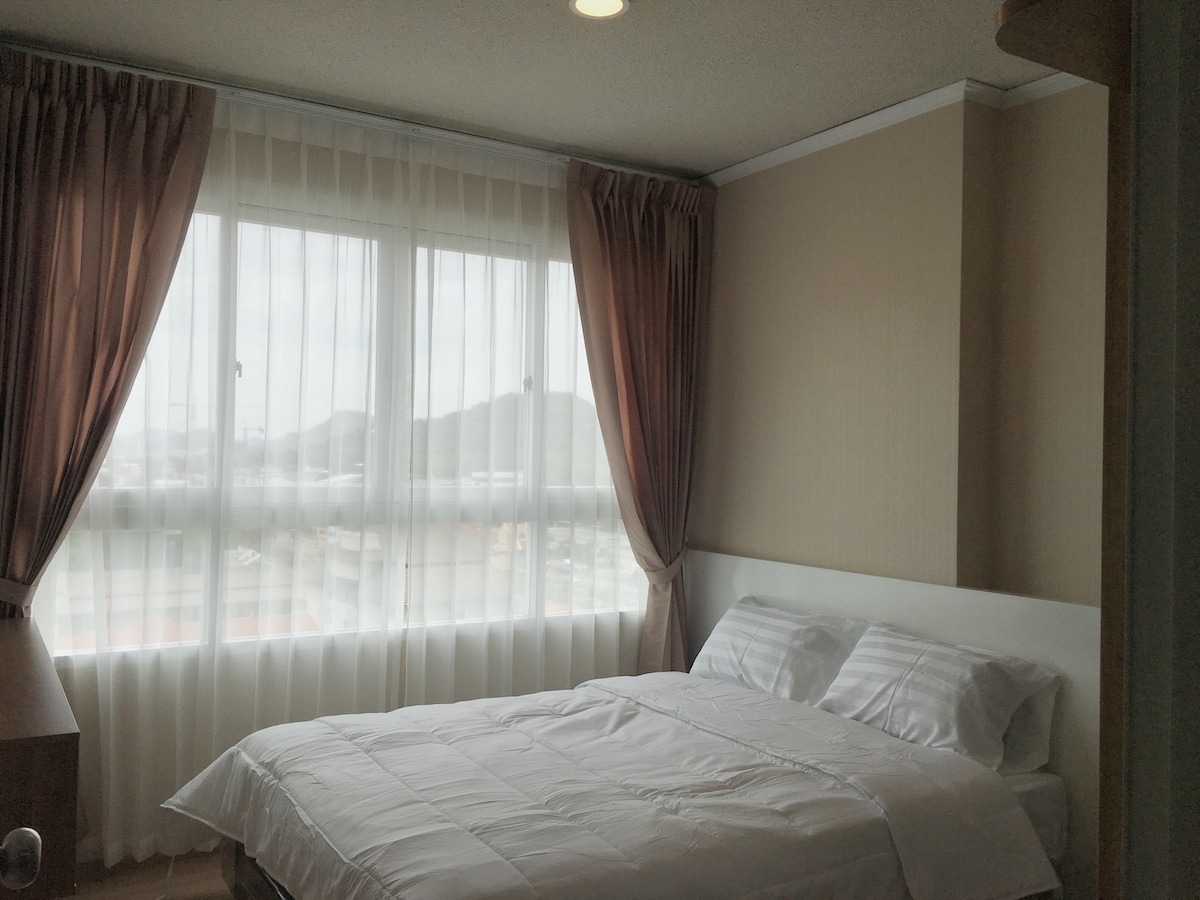 A well-lit bedroom features a spacious bed with crisp white linens. Large windows with sheer curtains allow natural light to filter in, offering a view of the outdoors. Soft drapery frames the windows, creating a comfortable and inviting ambiance.