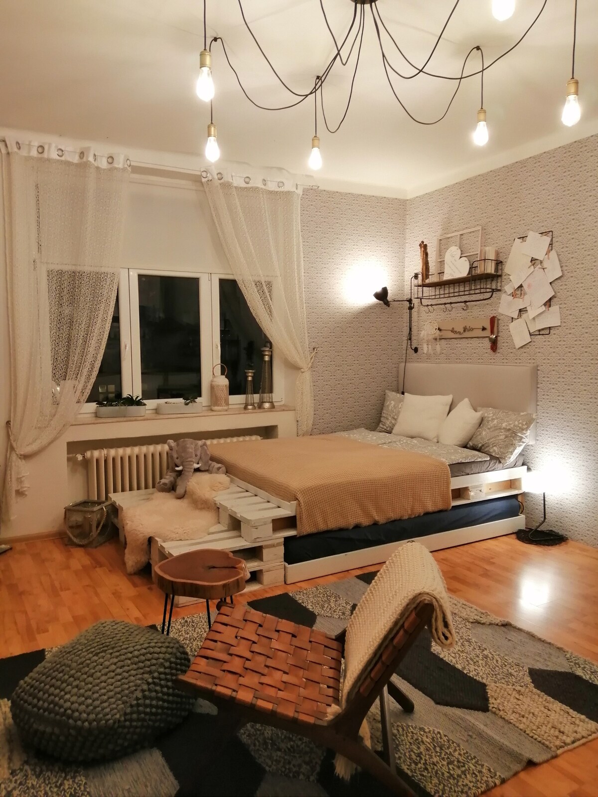 A comfortable sleeping area features a spacious bed dressed in warm bedding, accompanied by soft throw pillows. A window allows natural light to brighten the room, which is complemented by tasteful decor elements, including plants and wall art. A cozy seating area is created with a unique chair and textured textiles.