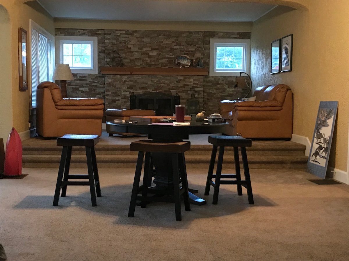 An inviting living area features two leather sofas positioned against a textured stone wall. In the foreground, three wooden stools surround a central table. Natural light filters through window panes, illuminating the carpeted space and creating a cozy atmosphere.