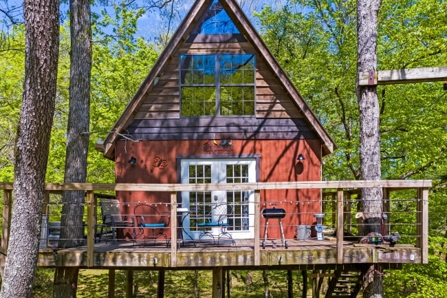 A charming treehouse is surrounded by lush greenery, featuring a wooden deck with seating and a grill. The triangular-shaped roof and large windows offer ample natural light, while the scenic view of the wooded lot enhances the peaceful atmosphere.