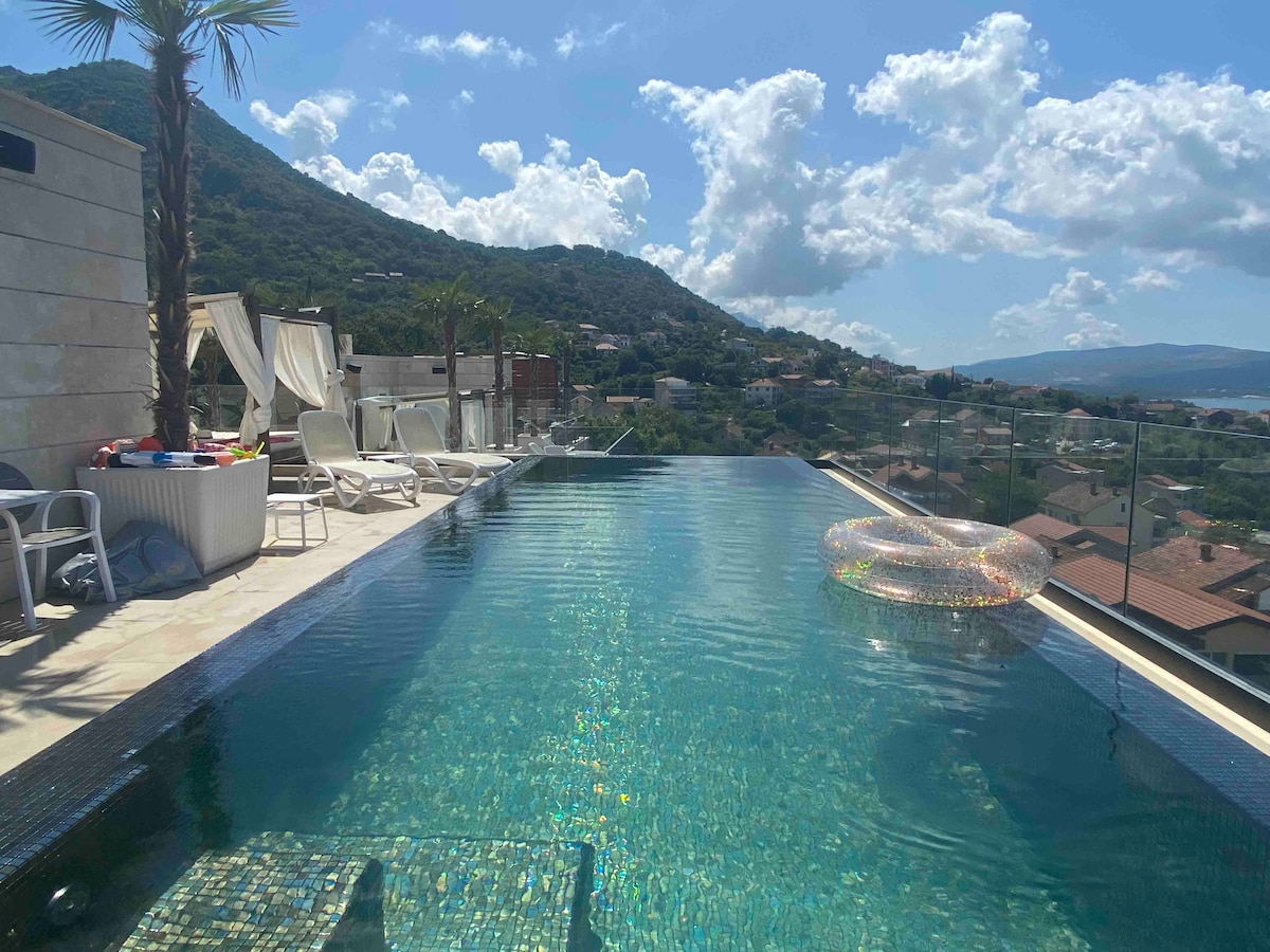 An infinity swimming pool stretches along the rooftop, reflecting the clear blue sky above. Lounge chairs are placed by the pool's edge, accompanied by a floatation ring. Lush greenery and hills can be observed in the background, creating a serene atmosphere.