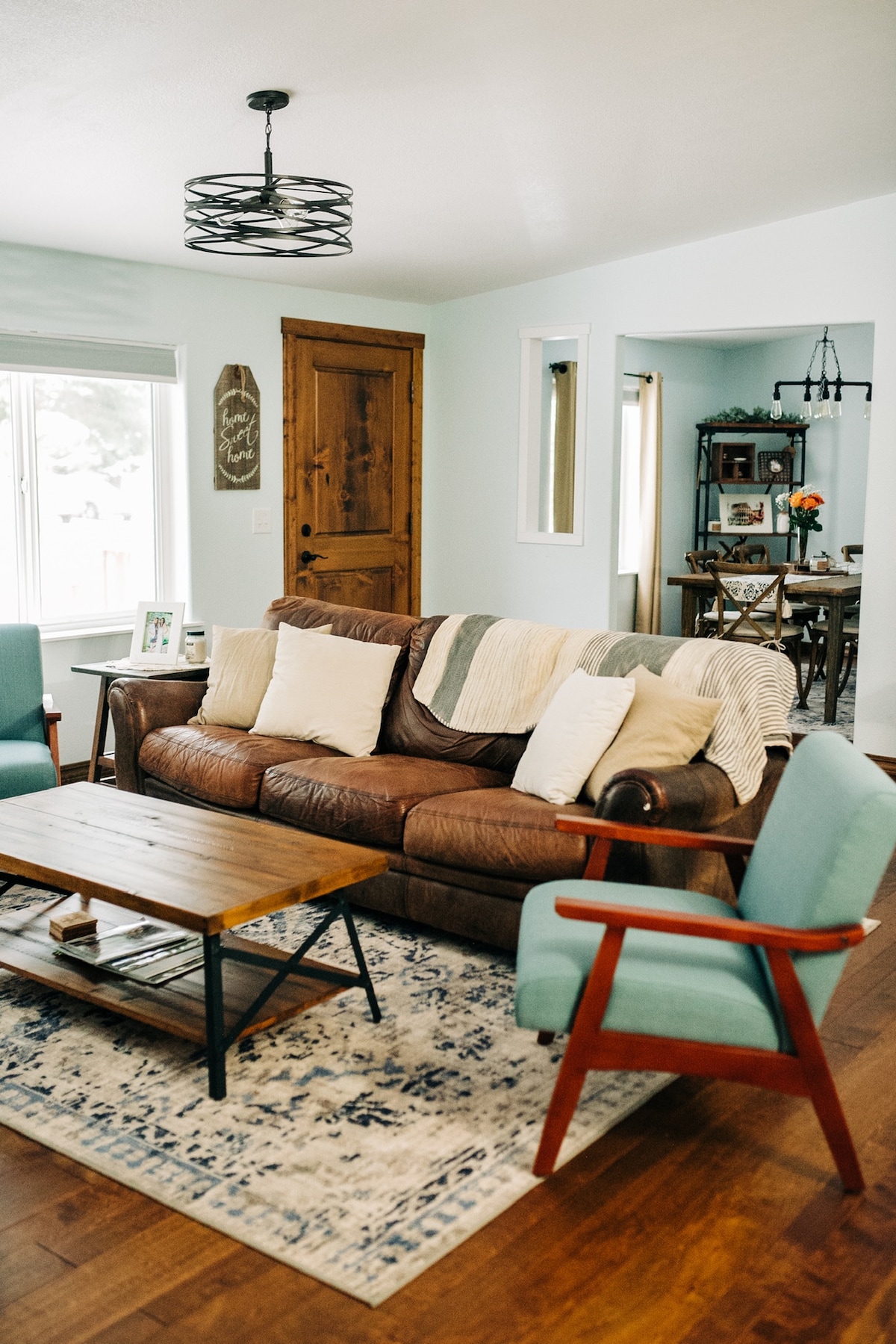 A cozy living area features a brown leather sofa adorned with multiple throw pillows. A wooden coffee table sits on a light blue and beige area rug. Two green accent chairs flank the coffee table, while natural light enters through the nearby window.
