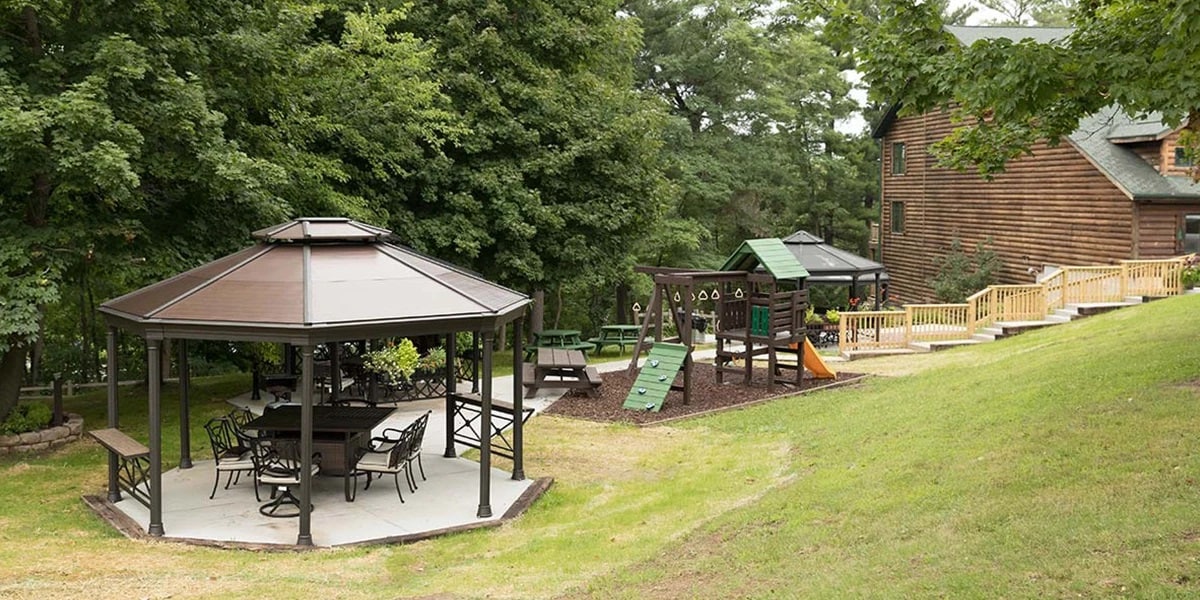 A spacious gazebo with multiple picnic tables is positioned on a grassy area, surrounded by trees. Adjacent is a playground structure, featuring slides and climbing elements, offering a fun space for outdoor activities. A wooden deck area is visible in the background.