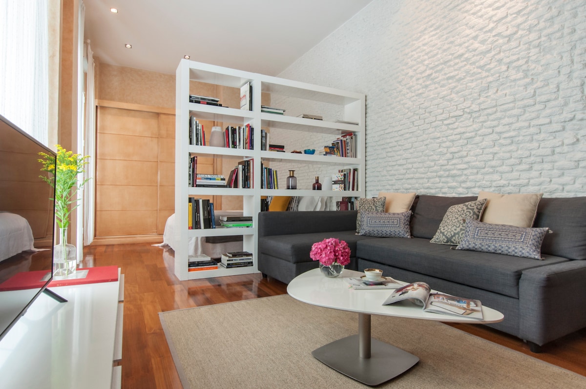 A modern living area features a cozy dark sofa with decorative cushions and an open bookshelf filled with books. A round coffee table sits in front, displaying magazines and a small vase of pink flowers. Natural light brightens the space through large windows.