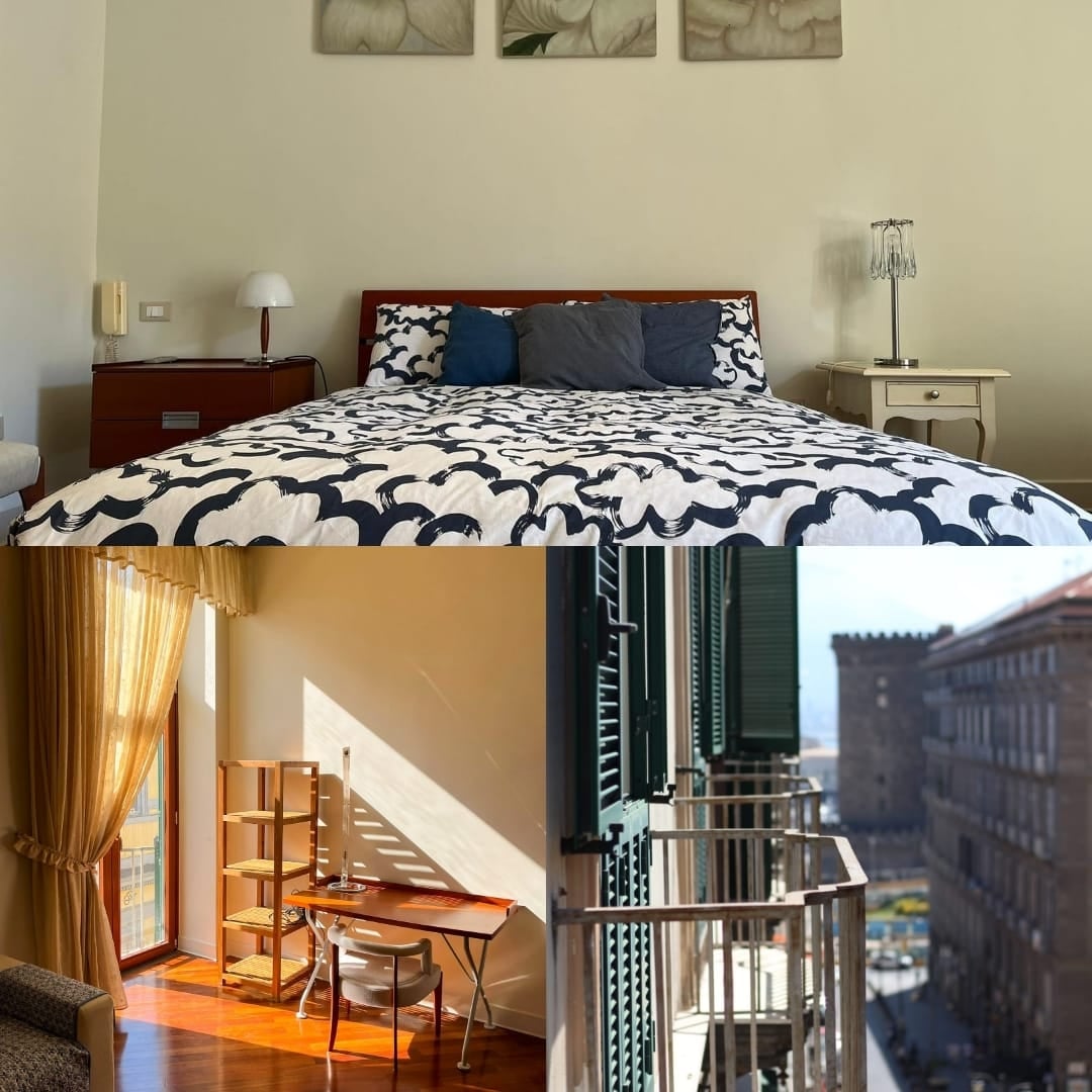 A spacious sleeping area features a patterned duvet covering the bed, complemented by two decorative pillows. Natural light is captured streaming through a window, illuminating a wooden desk and chair nearby, while a balcony showcases views of the vibrant street below.