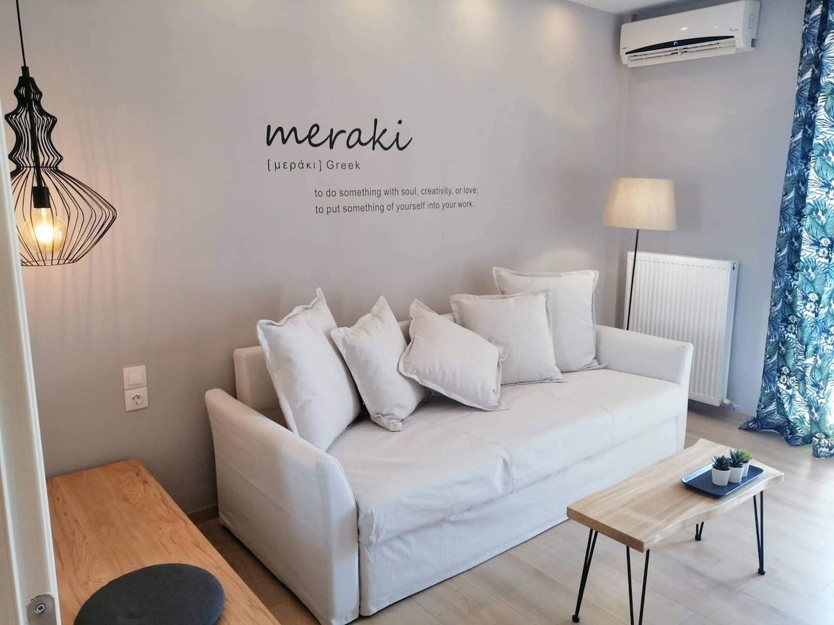 A living room area features a white sofa adorned with multiple soft, white pillows. A wooden coffee table sits in front of the sofa. Light from a decorative pendant lamp casts a warm glow, while patterned curtains frame a nearby window.