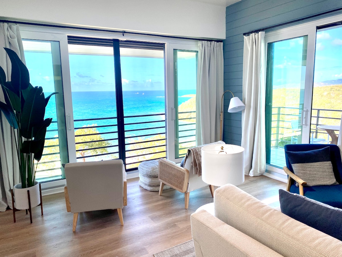 A bright living area showcases large glass doors with stunning ocean views, allowing natural light to illuminate the space. Comfortable seating options include a light-colored sofa and cozy armchairs. A decorative plant stands beside a clean white end table, contributing to the airy atmosphere.