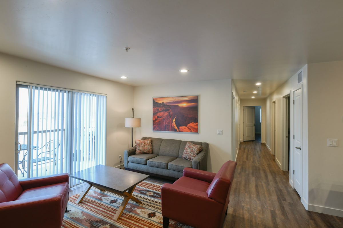 A living area is highlighted by a comfortable gray sofa with decorative pillows, flanked by two red armchairs. A wooden coffee table is centered on a patterned rug, with an artwork depicting a landscape on the wall. Natural light enters through sliding glass doors with vertical blinds.