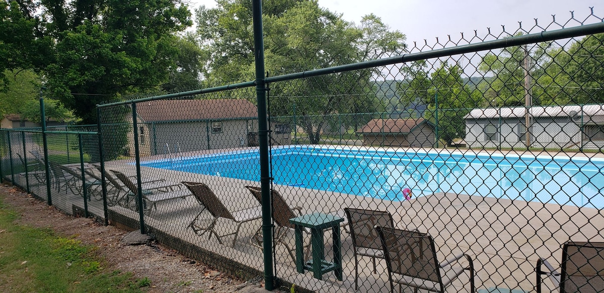 A fenced swimming pool area features a large pool with clear blue water, surrounded by a concrete deck. Several lounge chairs are positioned along the edge, offering a place to relax. Lush green trees and a few buildings are visible in the background.