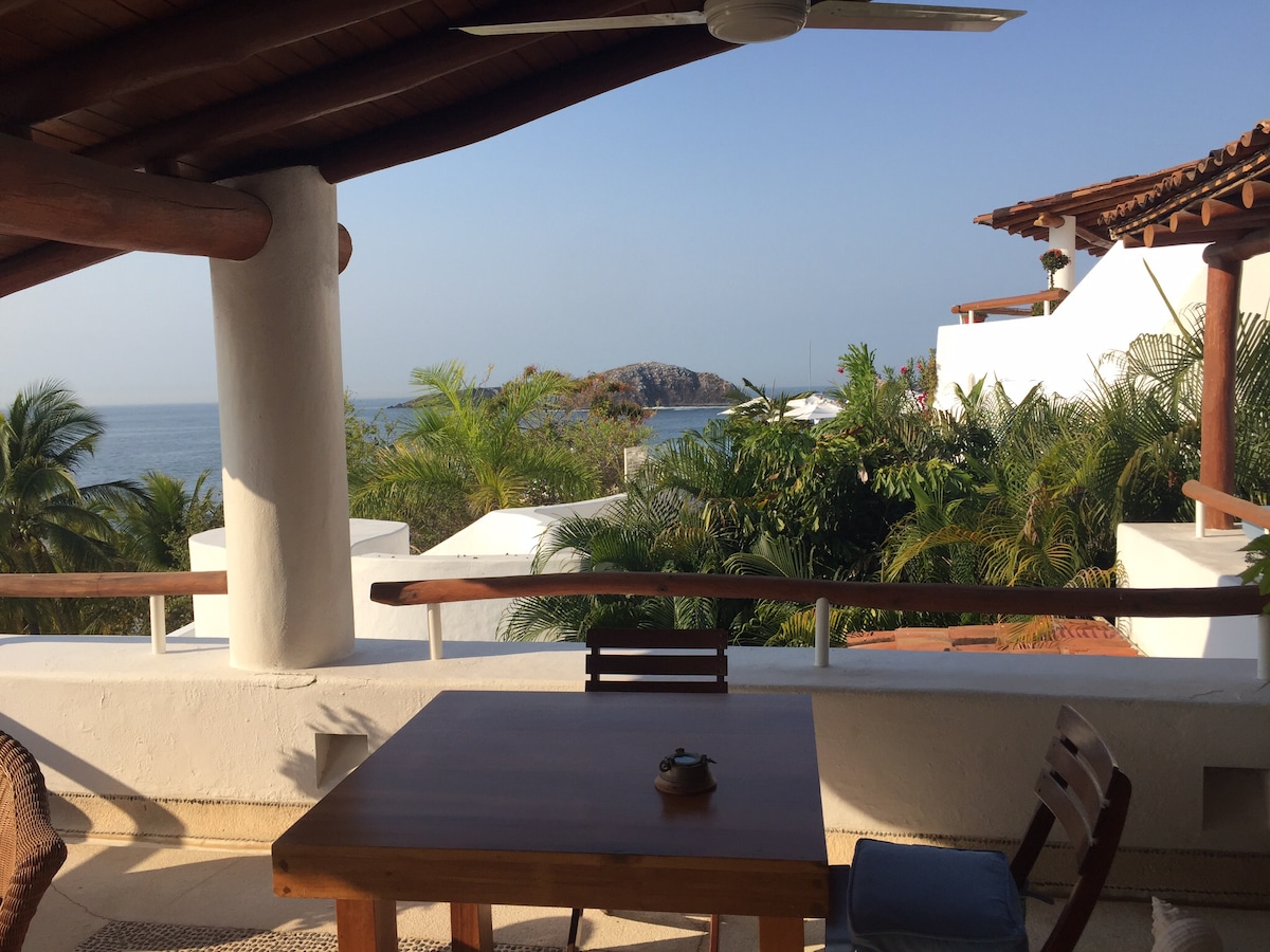An outdoor terrace is visible, featuring a wooden table with a small decorative item positioned atop. Surrounding palm trees and lush greenery frame the view of the Pacific Ocean, while distant rocky formations and white buildings complete the serene coastal setting.