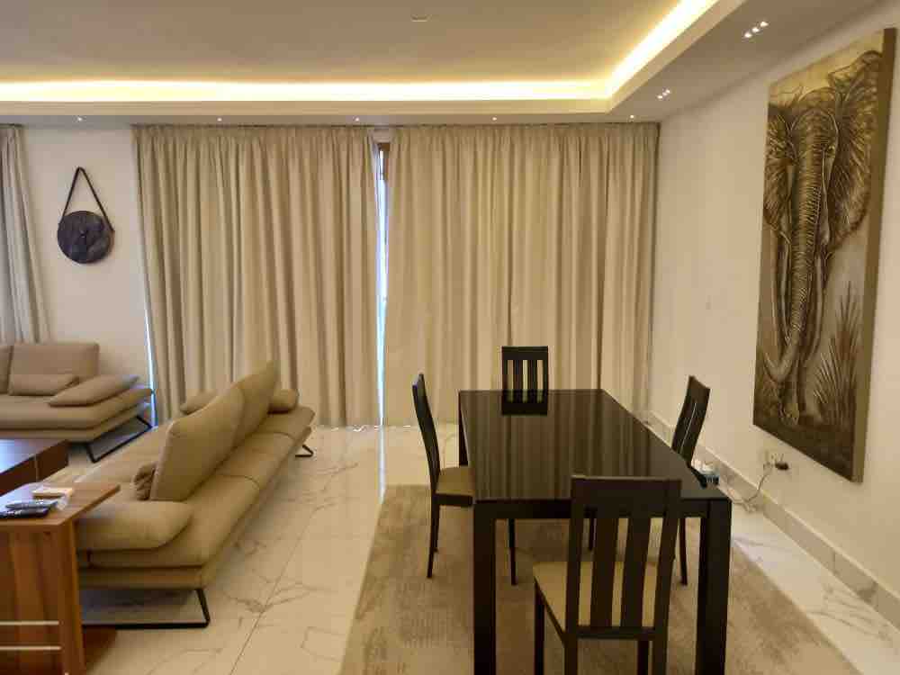 A dining area is presented with a sleek black table accompanied by four chairs. A light-colored sofa sits nearby, complementing the soft drapes that frame the windows. The room features a neutral color palette with a decorative wall art piece showcasing an elephant.