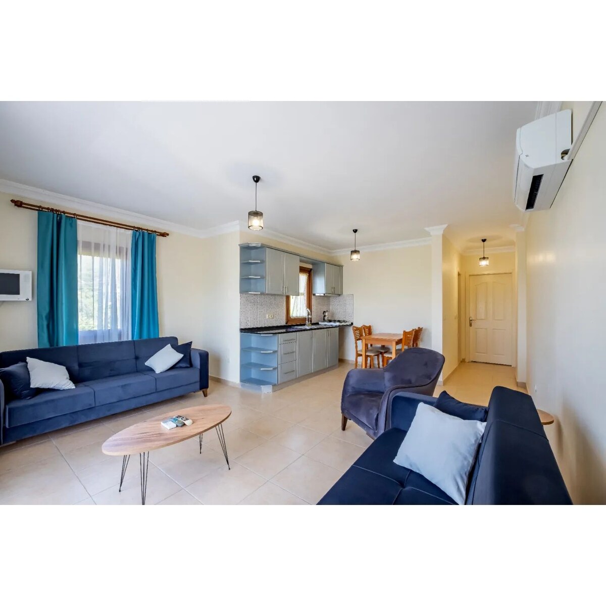 A living area features two navy sofas positioned around a small wooden coffee table. A dining table and chairs are visible in the background, adjacent to a kitchen with modern cabinetry. Large windows are adorned with blue curtains, allowing natural light to fill the space.
