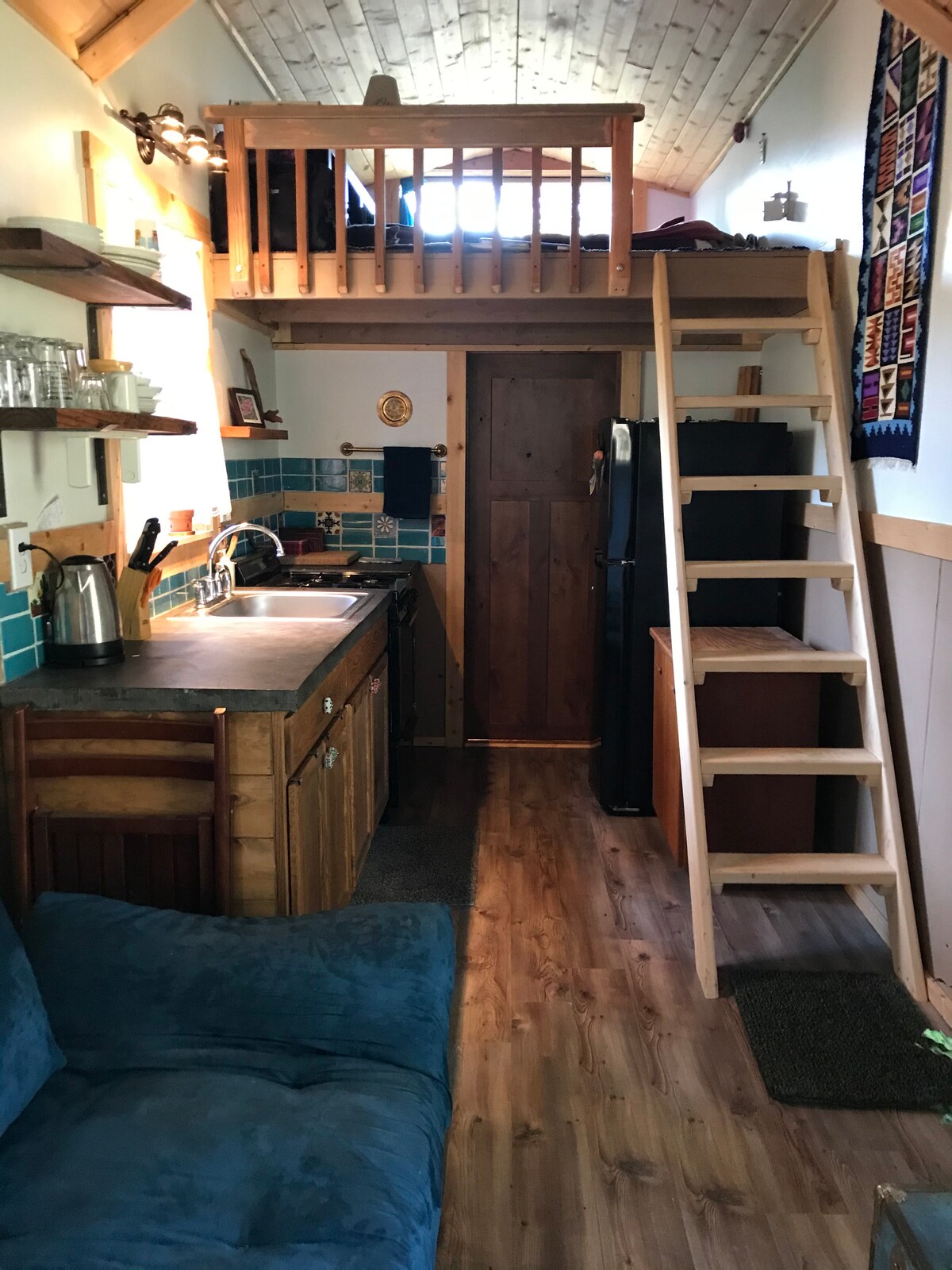 The interior of the tiny house features a compact kitchen area with wooden cabinetry and colorful backsplash tiles. A cozy seating area is present, along with a wooden ladder leading to a lofted sleeping space. Natural light enters through the windows, enhancing the inviting ambiance.