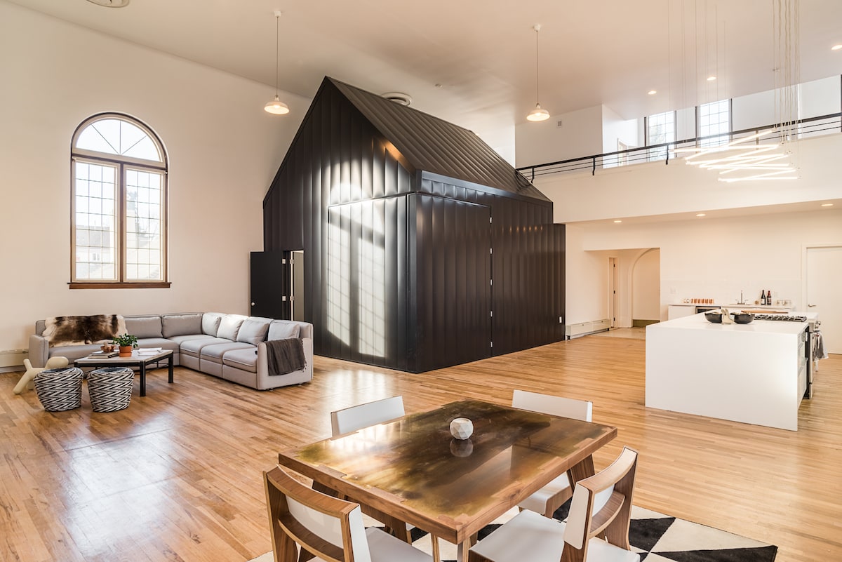 An open-concept living area features high ceilings and large windows, allowing ample natural light. A modern kitchen with a central island is visible alongside a dining table. A unique black structure, resembling a small house, adds architectural interest to the space.