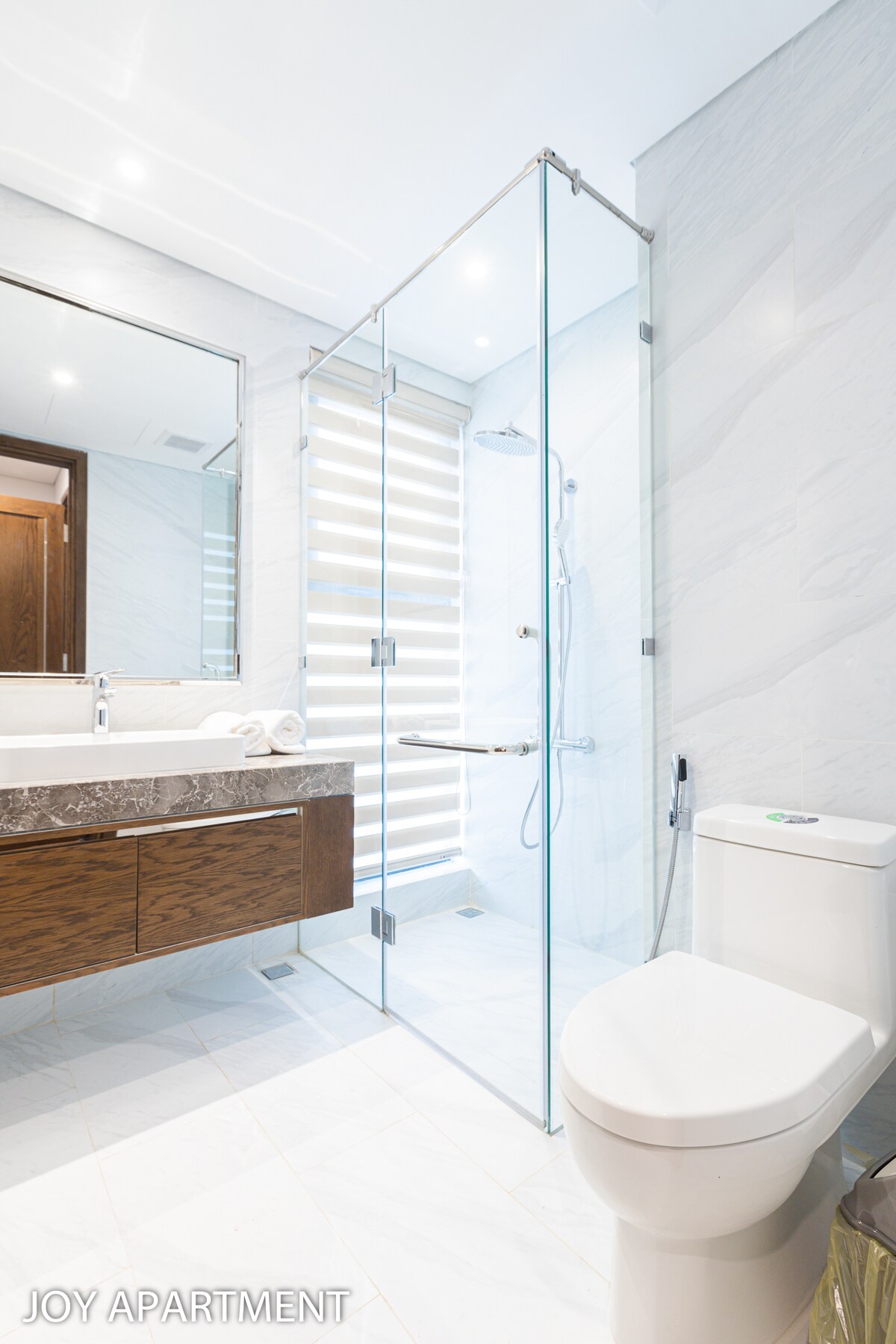 A modern bathroom is featured with a walk-in shower enclosed by clear glass doors. A sleek wooden vanity with a large sink is positioned beside the shower. Soft lighting illuminates the space, highlighting the neutral tones of the tiles and fixtures.