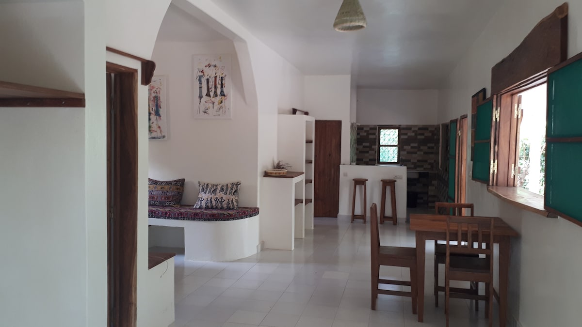 An open living area features a cozy seating nook with patterned cushions, complemented by a wooden dining table and chairs. Light-colored walls enhance the spacious feel, while large windows provide natural light and view of the garden outside.