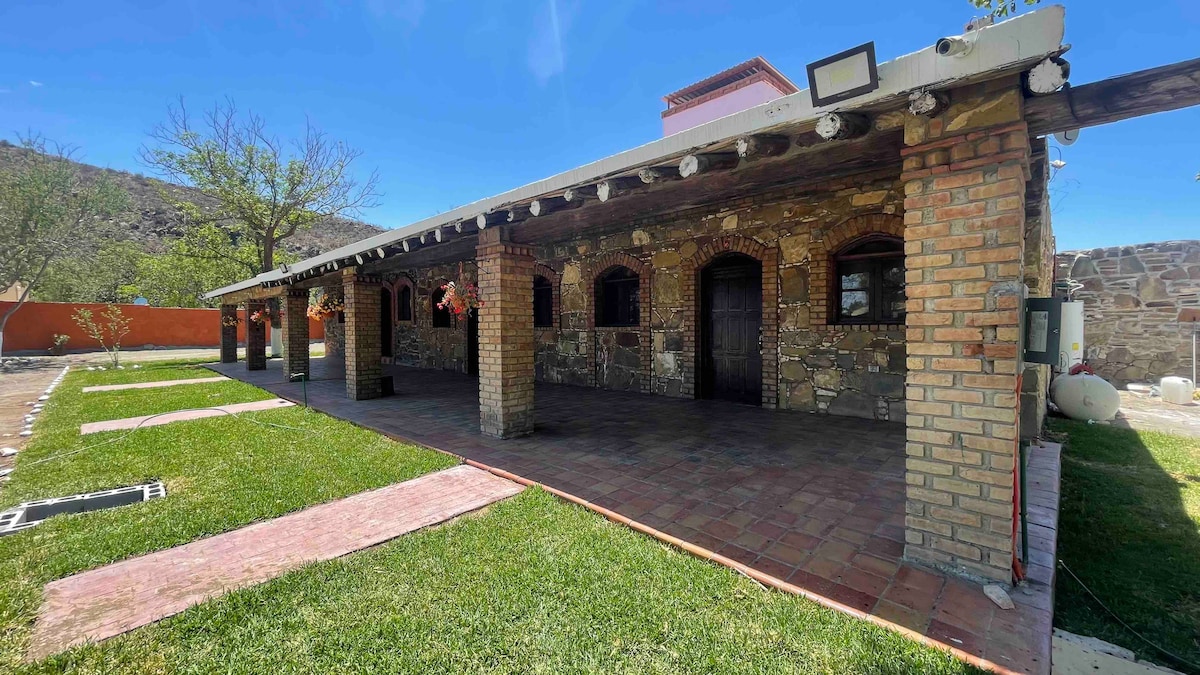 A charming stone facade is showcased, featuring a covered porch lined with wooden beams. The expansive outdoor area includes green grass and pathways made of bricks. Large windows provide views of the interior, with hanging flower baskets adding subtle color.