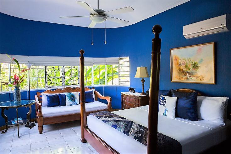 The room features a spacious king-size bed alongside a twin-sized daybed, both adorned with decorative pillows. Light filters through large windows, while a ceiling fan provides circulation. A painting and a small table with a lamp complete the inviting tropical ambiance.