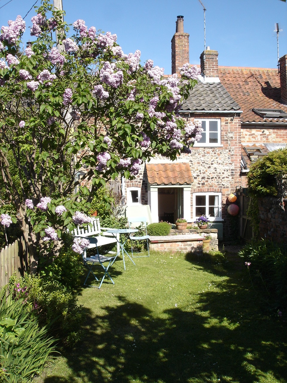 A charming garden features blooming lilac trees and green grass. A small patio area is seen with a table and chairs, providing a cozy outdoor seating option. The back of the cottage, with its traditional brick exterior, is framed by foliage, creating a peaceful retreat.