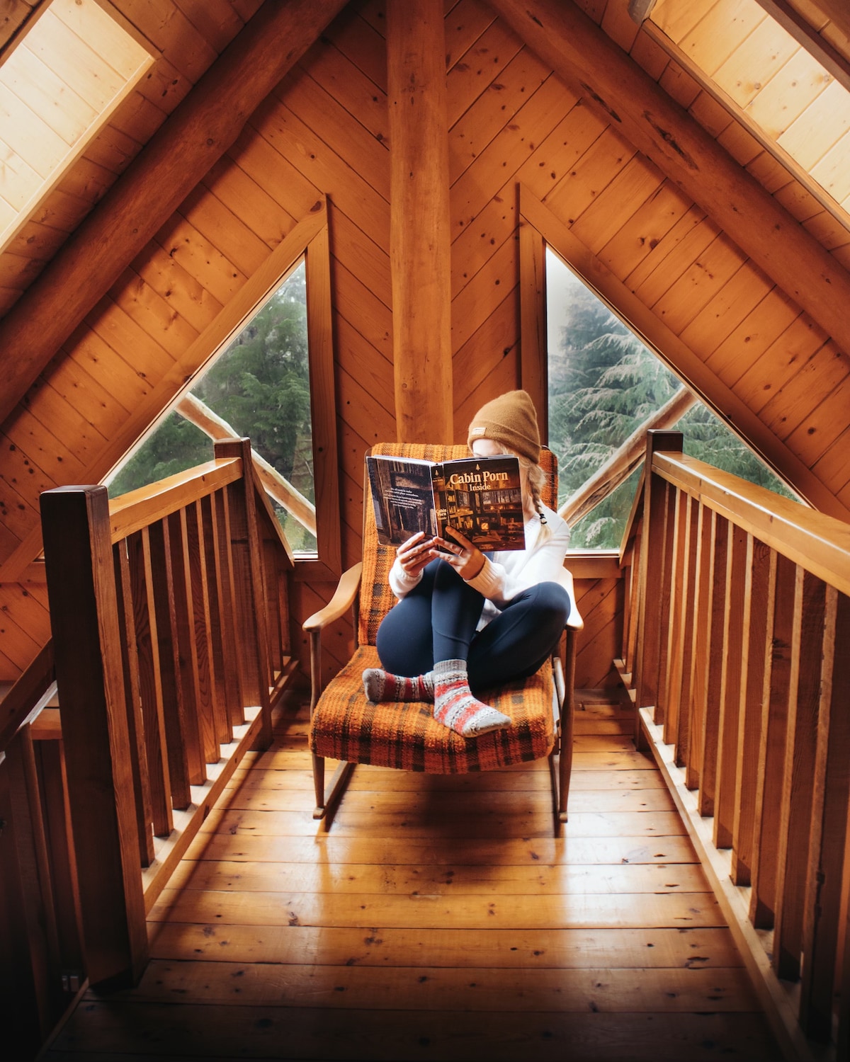 A cozy nook is depicted with a person seated in a plaid chair, engaged in reading. The space features wood paneled walls and triangular windows, allowing natural light to illuminate the area. Soft textures and warm tones create a serene reading atmosphere.
