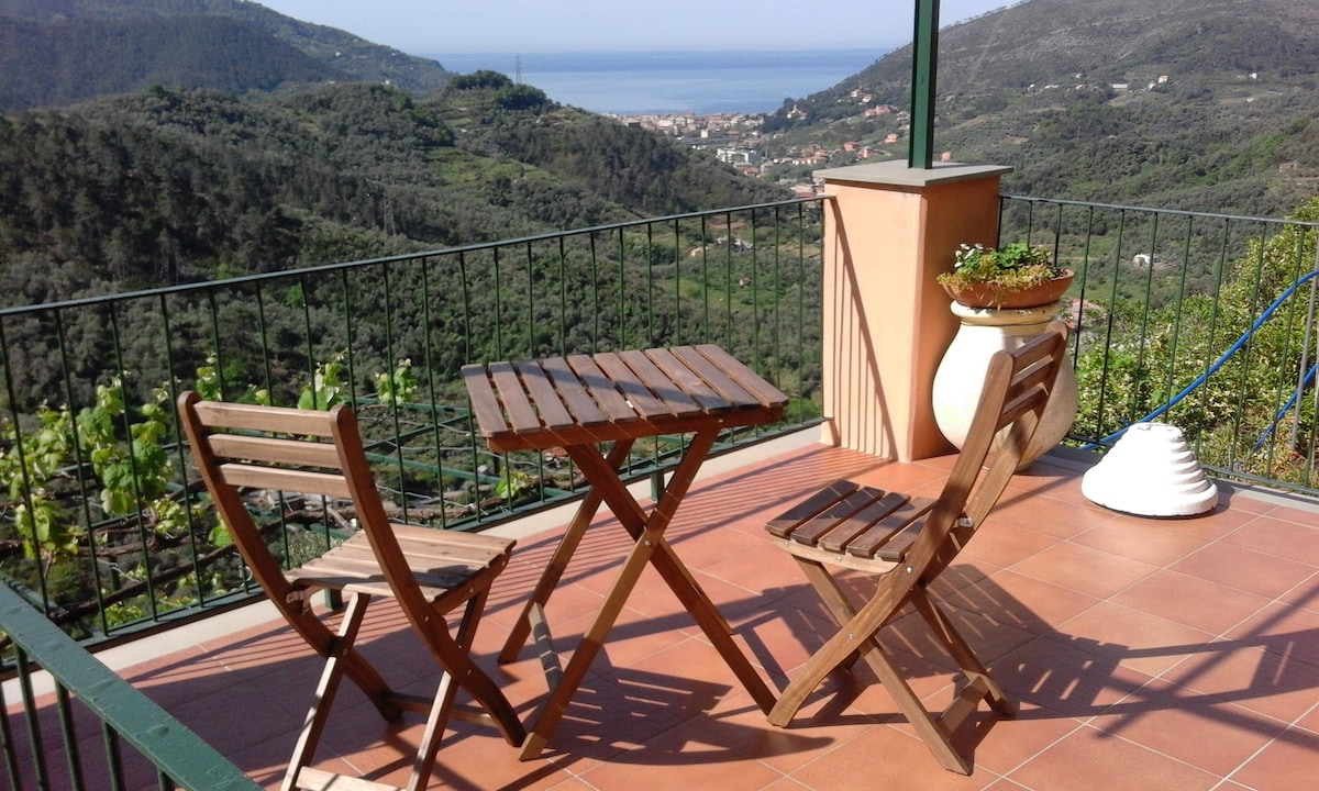 A sunny terrace features a small wooden table and two folding chairs, providing a space for outdoor seating. The surrounding landscape showcases lush hills and a distant view of the coastline, offering a tranquil setting to enjoy meals al fresco.