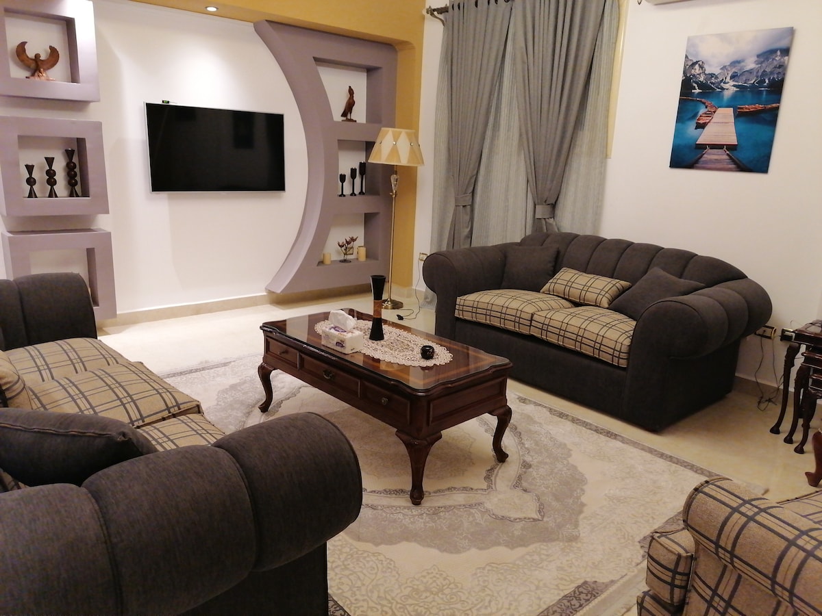 The living area features a comfortable gray sofa set arranged around a decorative coffee table. A flat-screen TV is mounted on the wall, complemented by elegant shelving displaying decorative items. A soft area rug lays beneath, and large curtains frame the windows.