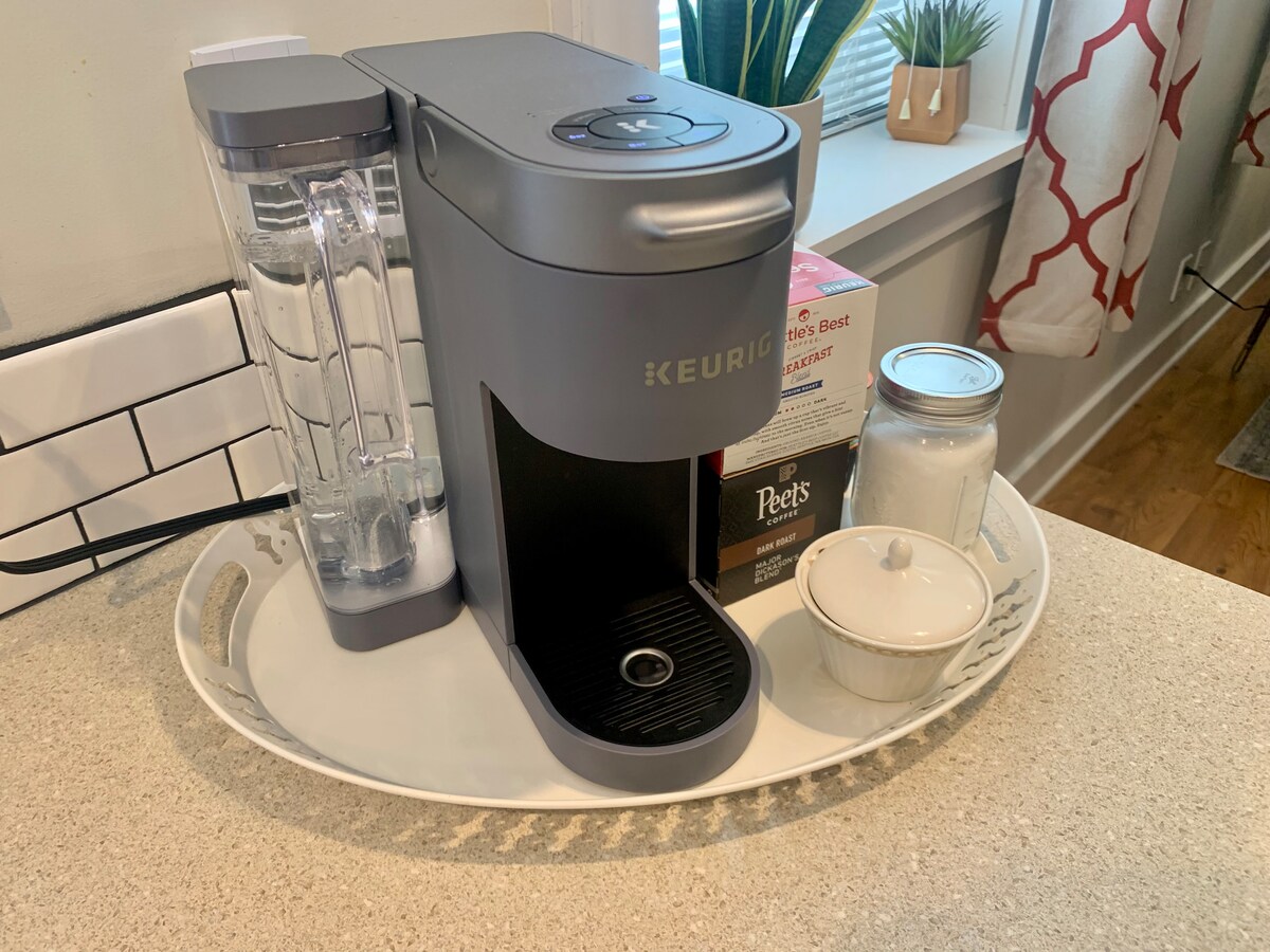 A Keurig coffee maker is positioned on a light-colored countertop, complemented by a clear water reservoir. Nearby, a decorative tray holds a box of coffee pods, a sugar bowl, and a glass jar, contributing to the functional and welcoming kitchen space.