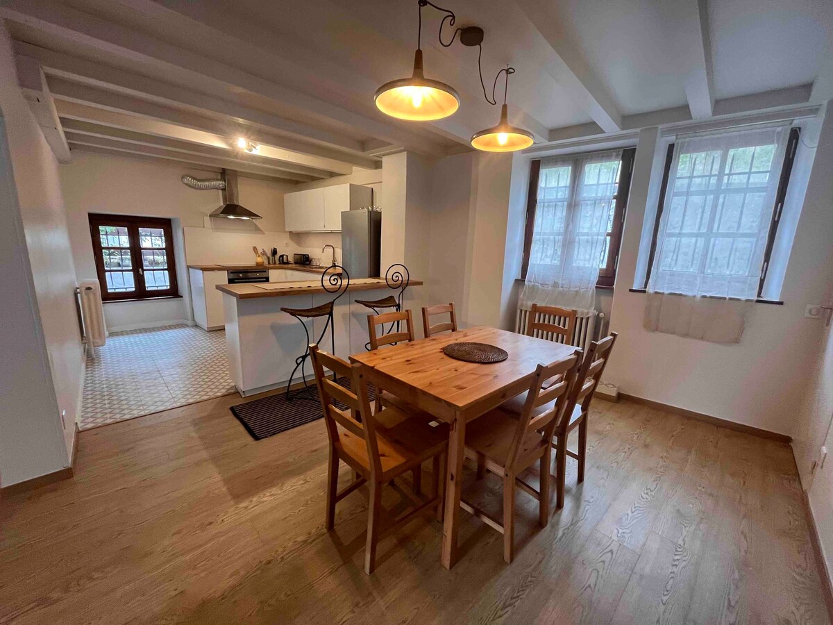 A spacious dining area features a wooden table surrounded by six matching chairs. Natural light filters through two large windows, enhancing the warm wood tones of the floors and furnishings. A modern kitchen with stainless steel appliances is visible in the background.