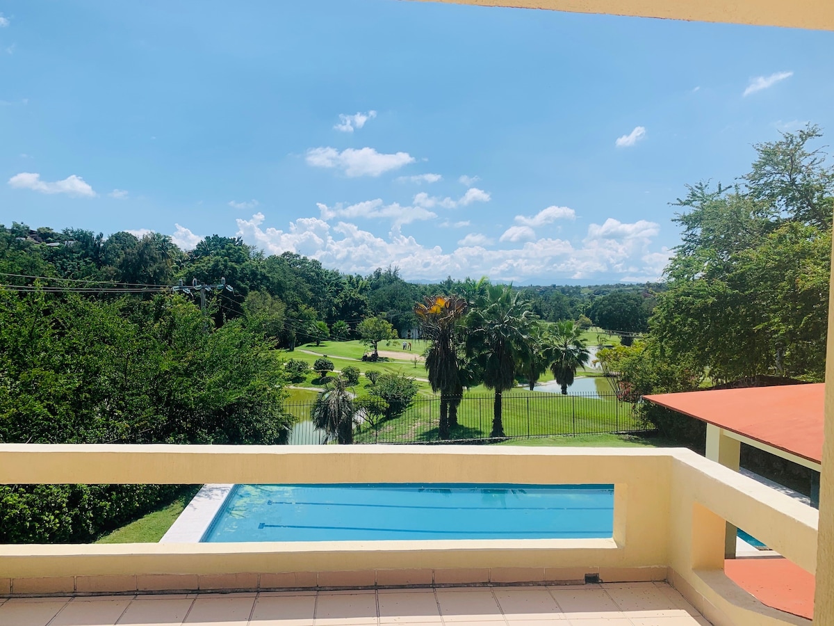 A serene view of a well-maintained golf course is captured from a terrace. Lush greenery, including palm trees, surrounds the area, complemented by blue skies and scattered clouds. The infinity-style pool is visible in the foreground, enhancing the tranquil setting.