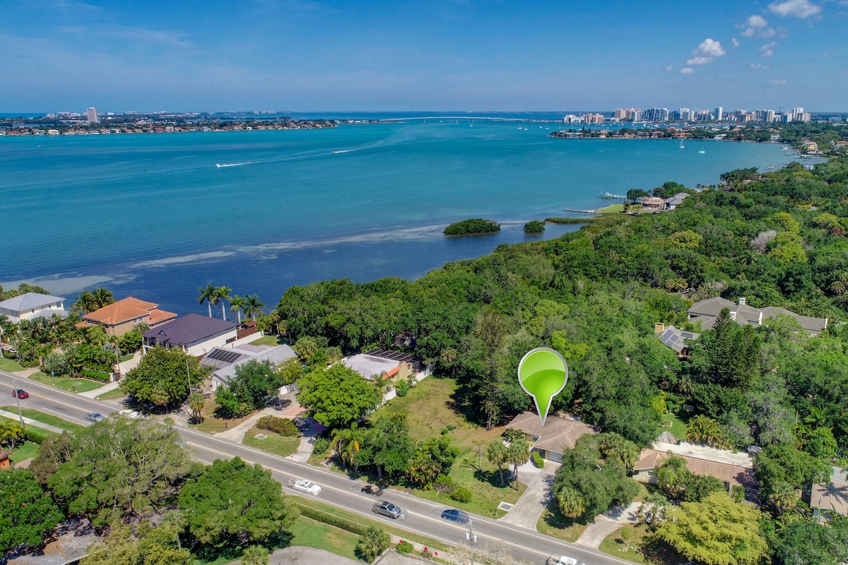 An aerial view captures a serene landscape featuring lush greenery and a calm blue waterway. Homes are visible along the shoreline, with distant city buildings punctuating the horizon. The inviting neighborhood is framed by clear skies and gentle waves, exemplifying a tranquil coastal environment.