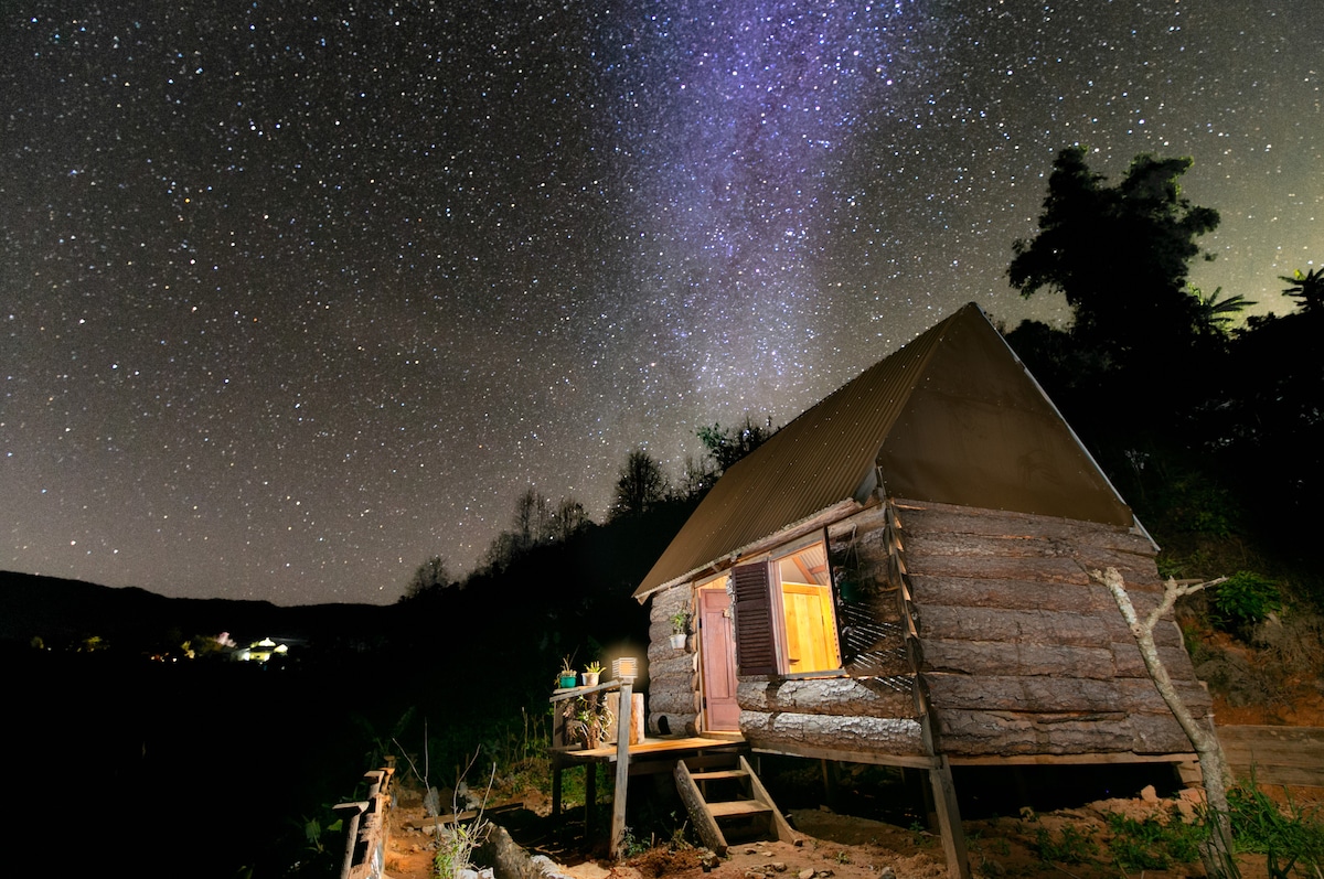 A wooden bungalow is illuminated by warm light, set against a clear night sky filled with stars. The surrounding landscape features distant trees and the faint outline of another structure. The peaceful scene invites quiet reflection and an appreciation of nature.