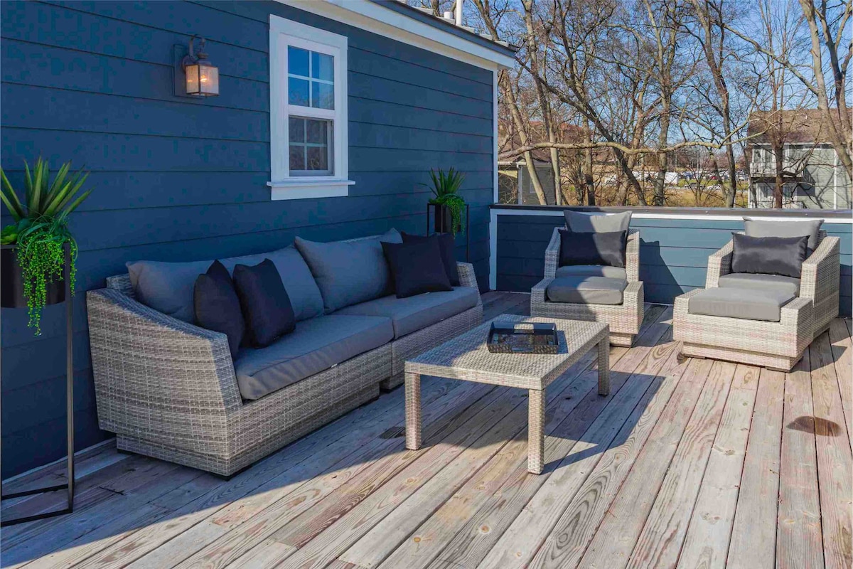 A spacious rooftop deck is presented with comfortable seating arrangements, including a sofa and two armchairs. A coffee table is centered on the wooden surface, while potted plants add a touch of greenery. The surrounding railing offers safety and unobstructed views of the outdoors.