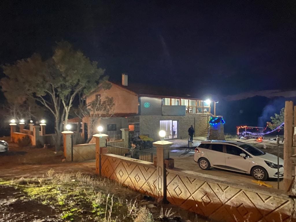 An evening view of a welcoming mountain house is observed, surrounded by softly glowing lights. A parked car is visible in the foreground, while trees frame the property, which features a spacious entrance highlighted by decorative lighting.