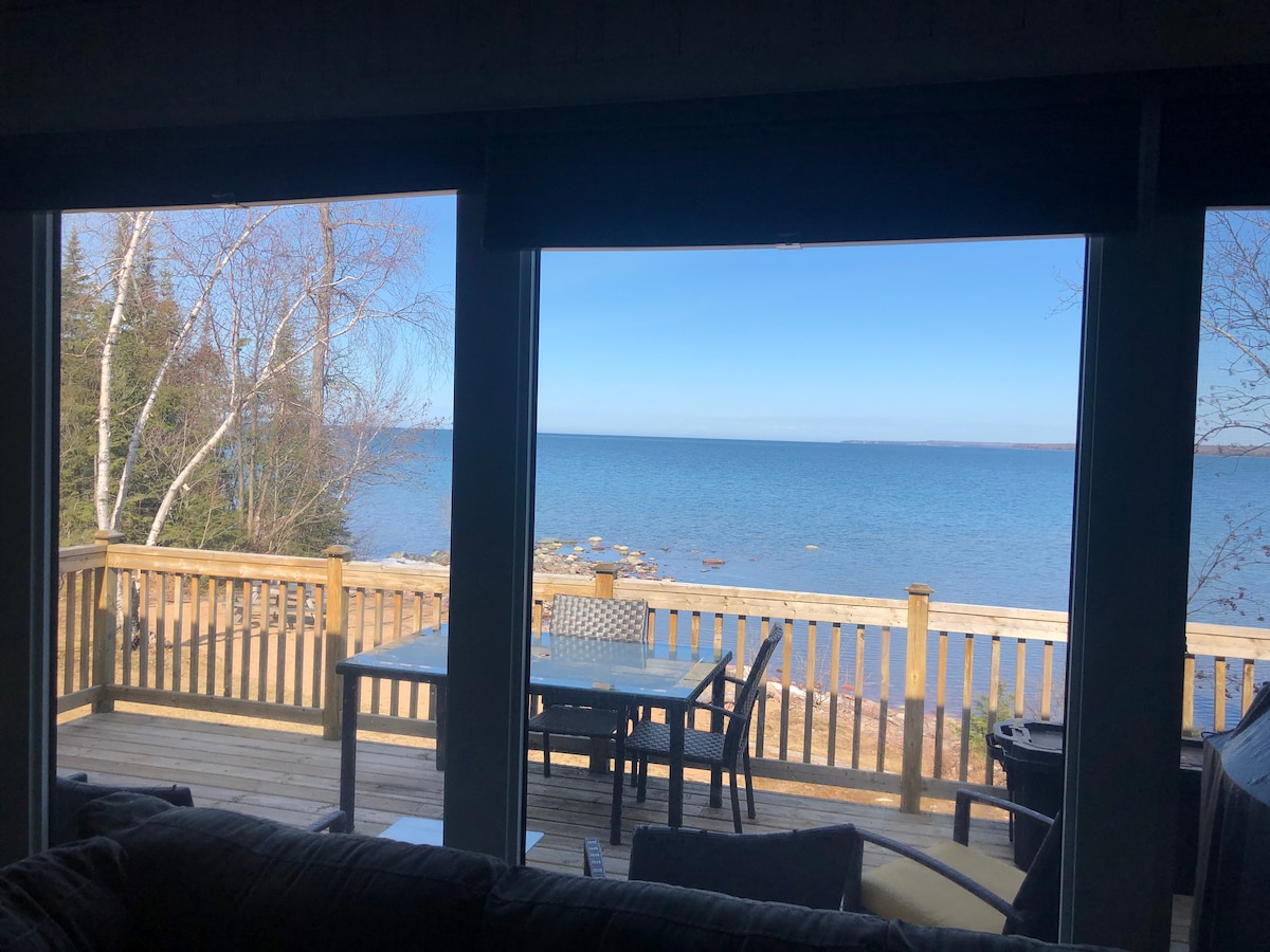 Large windows frame a scenic view of Lake Superior, showcasing calm waters and a sunlit shoreline. A wooden deck features a dining table and chairs, inviting outdoor relaxation. In the foreground, the plush seating area offers a comfortable vantage point to enjoy the natural beauty.