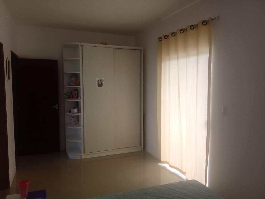 A bright and airy sleeping area features a spacious bed with a light-colored coverlet. A white wardrobe is positioned against one wall, and a tall shelf holds decorative items. Natural light filters through a soft curtain, enhancing the serene ambiance.