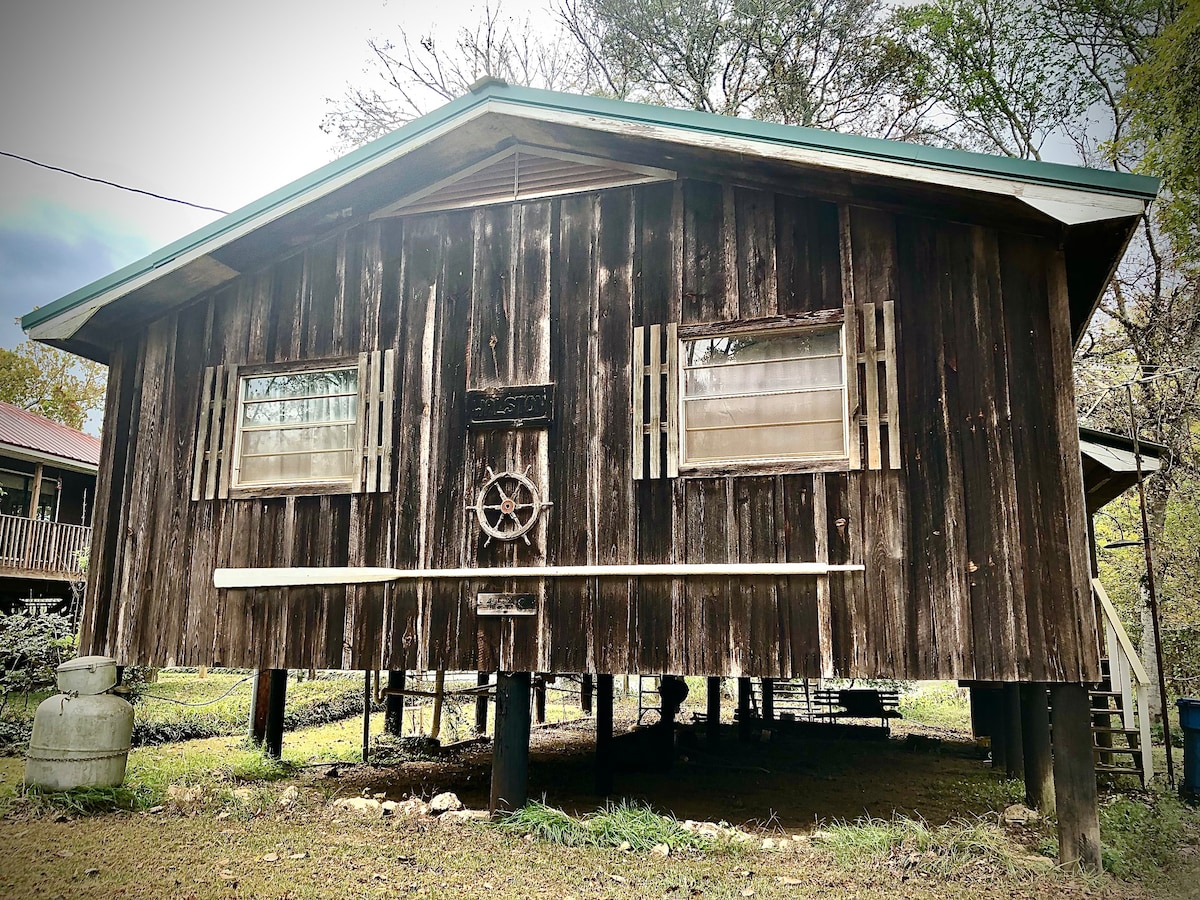 The exterior of the river house is characterized by weathered wooden panels and a peaked roof. Decorative shutters frame two large windows, while a ship's wheel adds nautical flair. The structure stands on wooden stilts, surrounded by a green lawn and trees.
