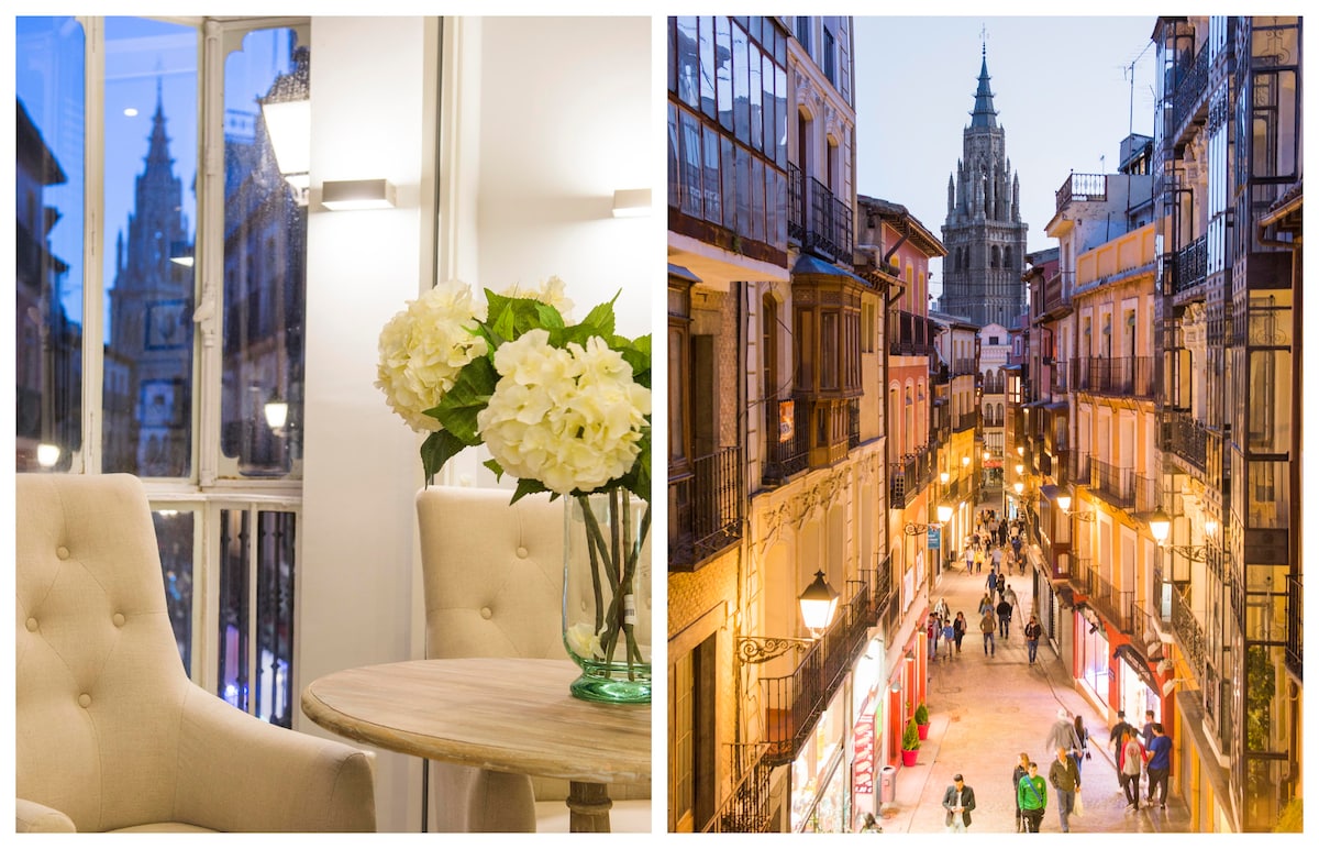 A cozy seating area features soft beige chairs positioned near a round wooden table. A vase of white flowers adds a touch of elegance to the interior. On the right, a vibrant street view shows people strolling past shops and restaurants, with the Toledo Cathedral visible in the distance.