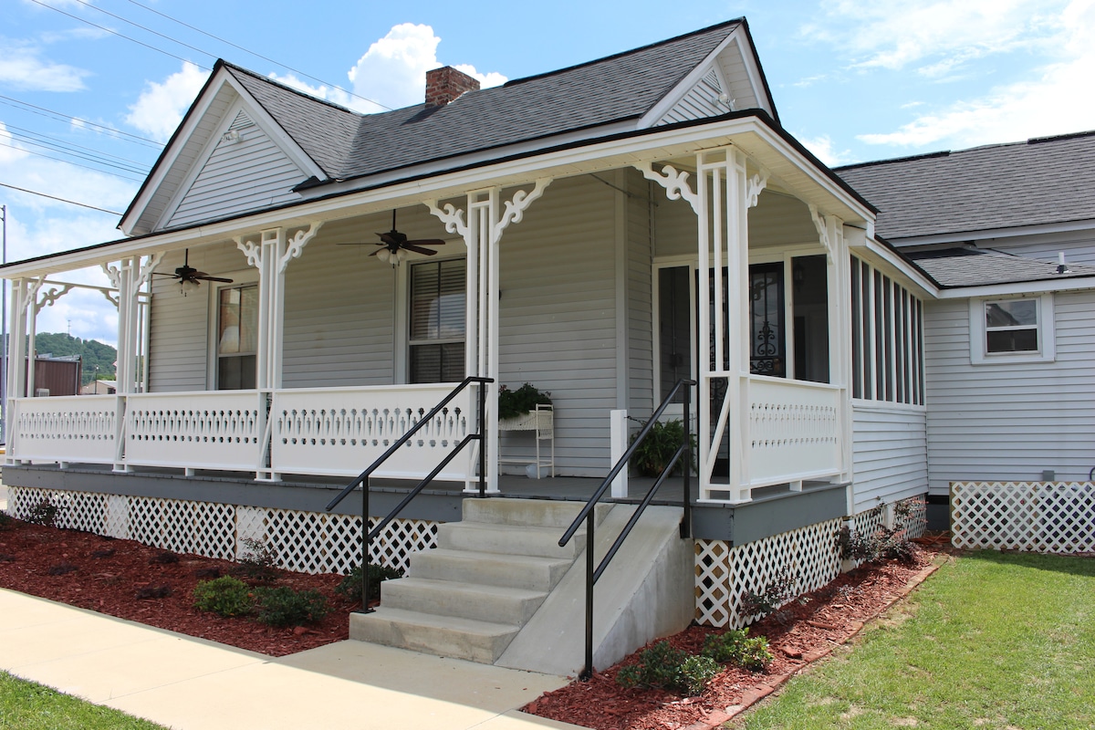The exterior of the Roberts Home Museum features a welcoming porch with decorative railings and ceiling fans. Steps lead to the entrance, which is framed by landscaping and a concrete walkway, set against a backdrop of clear blue skies.