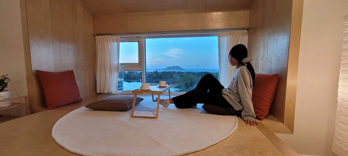 A cozy, circular seating area is positioned by a large window, showcasing views of the ocean and distant island. Soft cushions in warm tones are arranged for comfort. Natural light from the window creates a calming ambiance, enhancing the serene atmosphere.