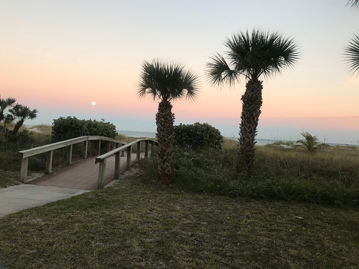 A wooden walkway leads towards the shimmering ocean, framed by palm trees with lush greenery on either side. The sky displays soft pastel hues of pink and orange, with the sun setting gently over the horizon.