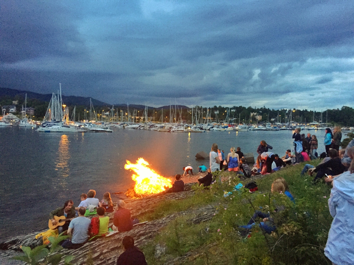 A lively gathering takes place near the waterfront, where people are seated on the rocks. A bonfire provides warmth, with flames visible against a backdrop of boats anchored in a marina. The sky is overcast, hinting at an approaching evening.