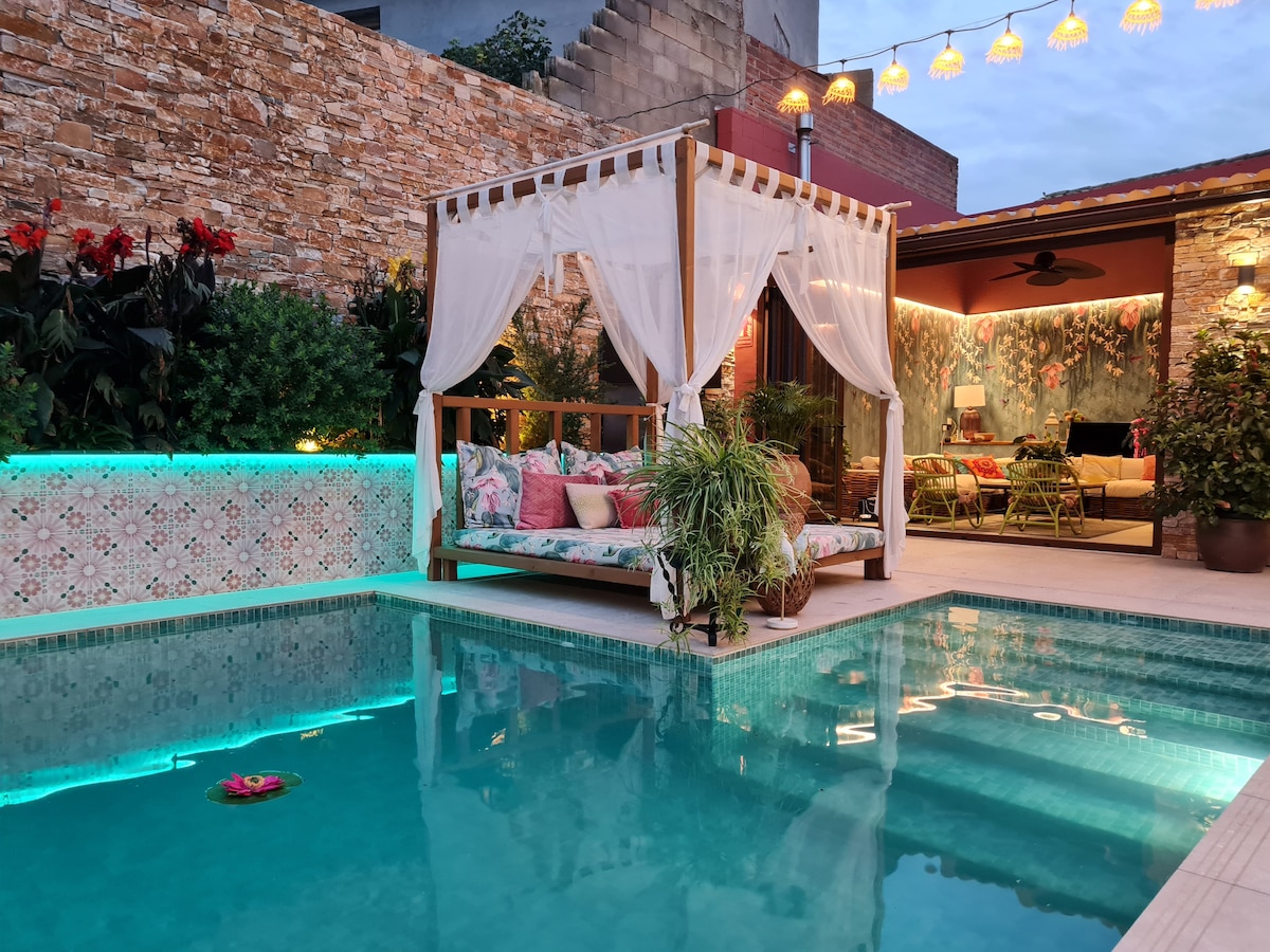 A private pool, framed by elegant tiles, reflects the evening sky. Nearby, a lounge area with a draped canopy is adorned with colorful cushions and greenery, providing a cozy retreat. Soft lighting from the surrounding decor creates a calm ambiance.