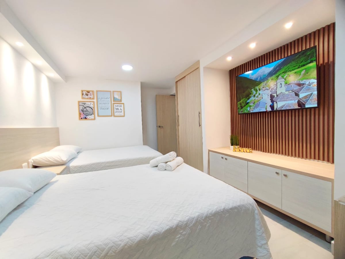 A modern sleeping area is equipped with two spacious beds, each topped with crisp white linens. A wooden television console holds a large screen, while art pieces adorn the walls. Soft lighting highlights the room's contemporary design and inviting atmosphere.
