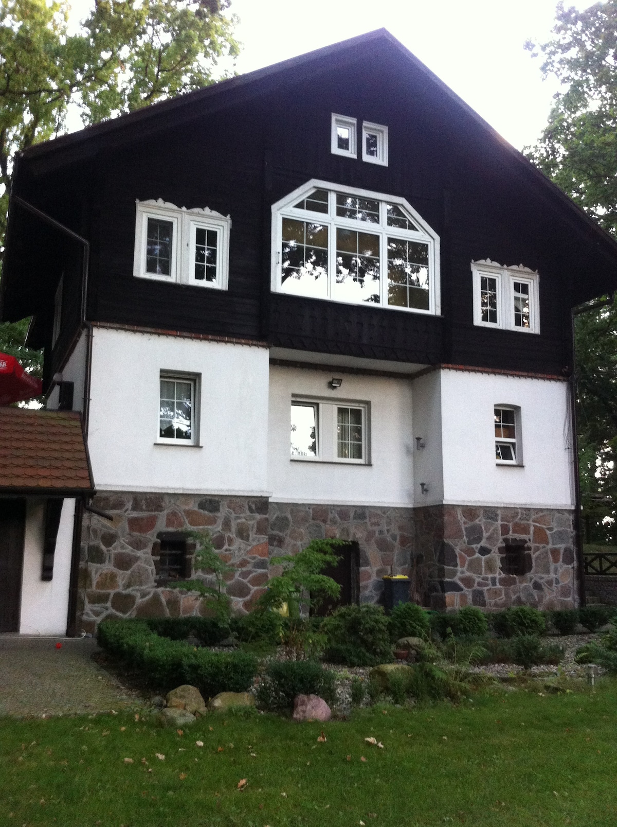 The exterior of a historic house features a combination of dark wood and stone materials. Large windows provide natural light, while a covered entrance and surrounding greenery add charm. A landscaped garden with stones and plants enhances the inviting appearance of the property.