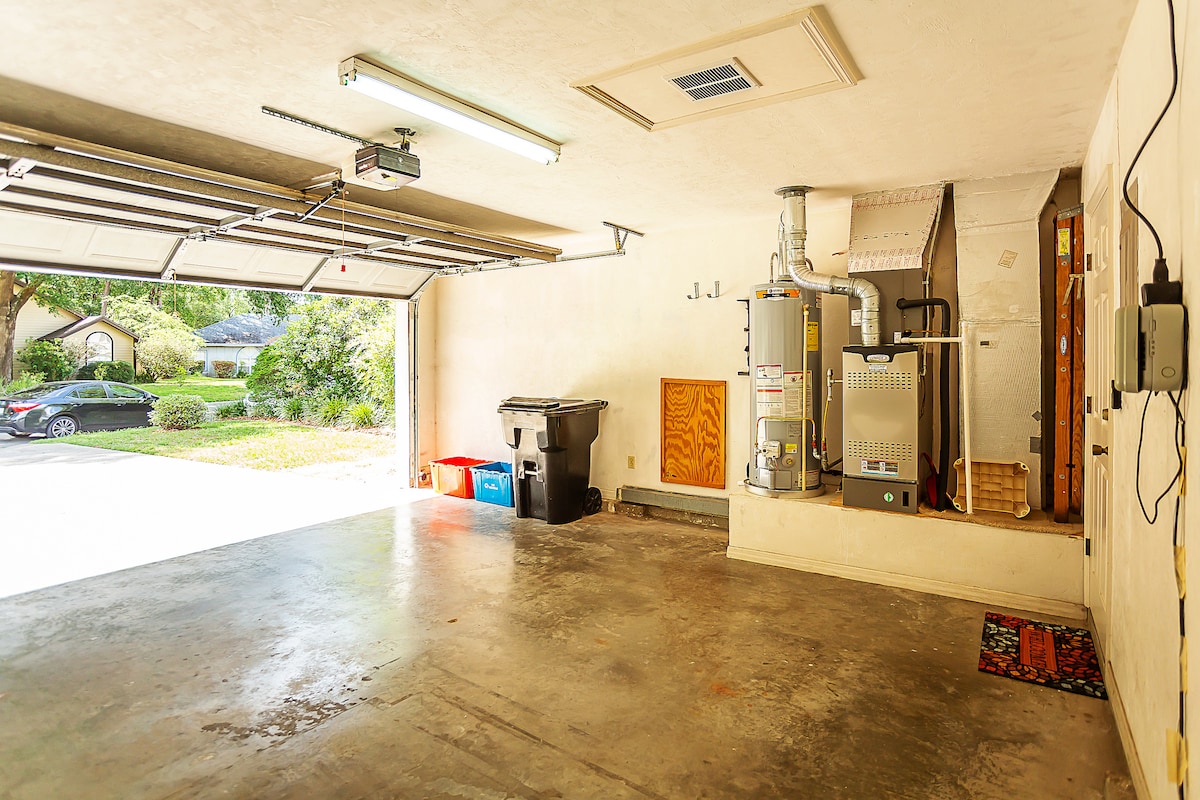 A spacious garage is presented with a concrete floor and abundant natural light from the open door. Visible items include a trash bin and a water heater. The garage walls are light-colored, enhancing the open feel of the space.